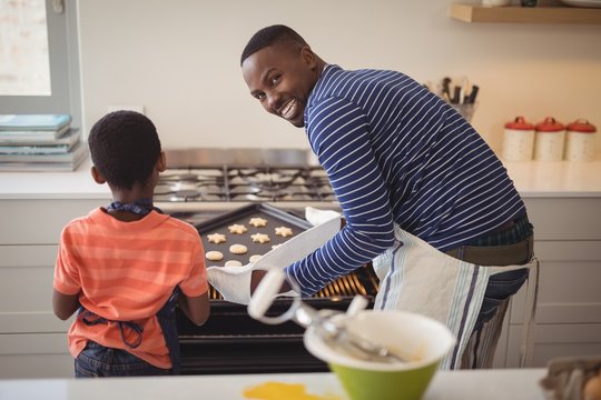Father taking tray of fresh cookies out of oven with son in