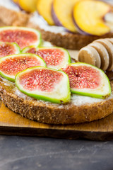 Toasts sandwiches with wholemeal rye bran bread, cream cheese or yoghurt, ripe figs and peaches. Wood honey dipper on cutting board. Close up.