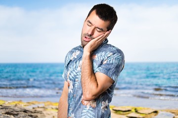 Handsome man with flower shirt making sleep gesture at the beach