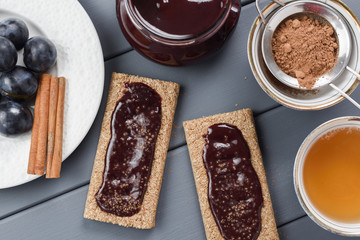Homemade plum jam on rye crackers served with cocoa, cinnamon, tea and fresh plums on gray table