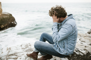 young man in denim jacket sitting by light blue ocean on white rocks