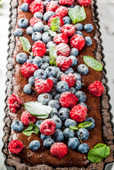 Summer homemade baked pastry. Chocolate cake tart with chocolate cream, fresh raw berries blueberry raspberry, decorated with mint leaves, powdered sugar. On white marble table, copy space close view
