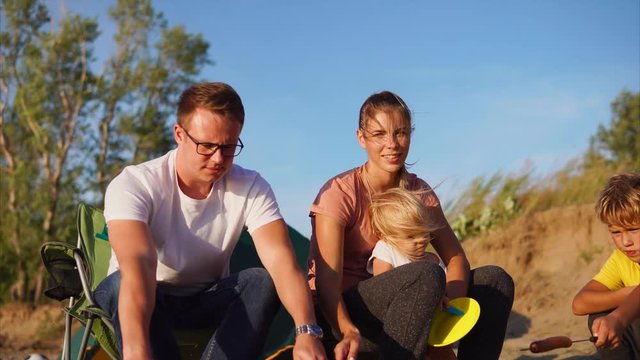 A young and happy family who rests on a sandy beach with fried meat sausages on fire, mom holds her little child, next to her son and husband sitting, who cooks dinner