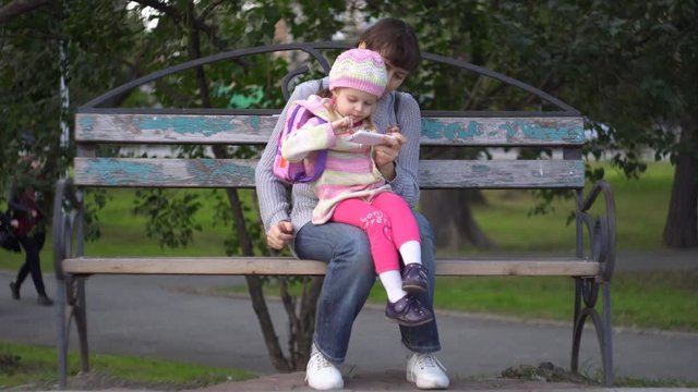 Mother with her little daughter dressed in a striped sweater together use the phone sitting on a bench in a public park on a cool evening. A young woman holds a baby girl on her lap.