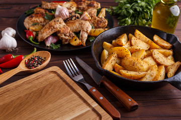 Grilled chicken wings, baked potatoes, herbs and spices on kitchen table. Hot fresh appetizing countryside food. Cooking concept, meat dishes, homemade cuisine