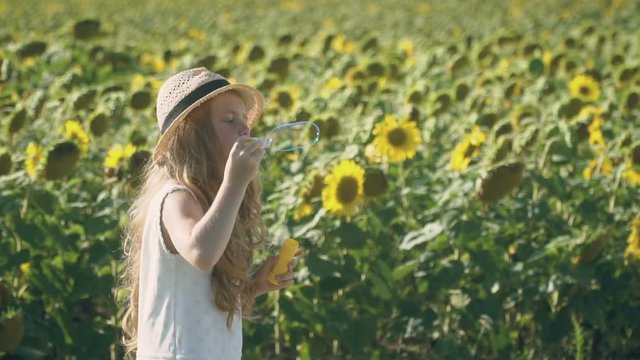 Pretty little girl in hat blows bubbles at sunflower field