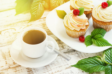 Delicate vanilla cupcakes with cream and raspberries on a white wooden background with a cup of coffee