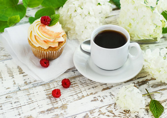 Delicate vanilla cupcakes with cream and raspberries on a white wooden background with a cup of coffee