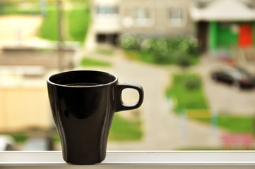 Black cup of coffee on the windowsill on a blurred city background.