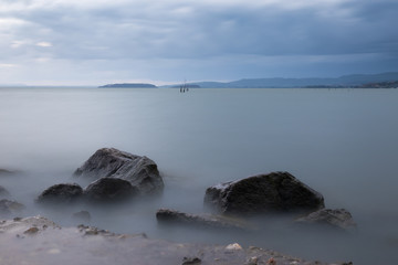 A minimalist, long exposure photo of some big rocks in a lake, with perfectly still water and cloudy sky