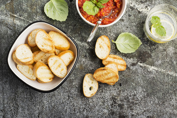 Healthy snack or lunch. Homemade tomato vegetable sauce with garlic, herbs, salt, butter, spices, fresh basil in a ceramic bowl with grilled toasts on a gray stone background. Top View.
