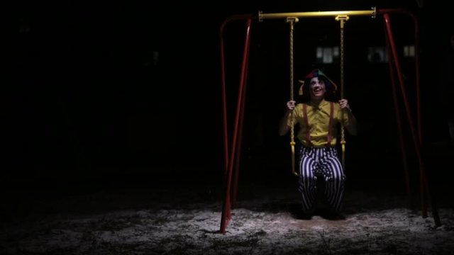 A clown rides low children swings in the dark. 