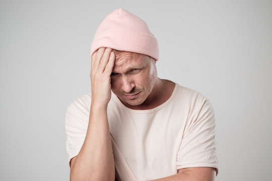 Portrait Of Sad Caucasian Man In Pink Hat. He Holds Head Because Of Pain And Looks Down.