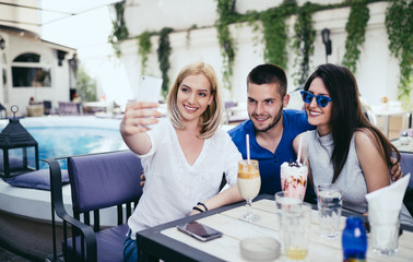 Group of young and attractive three people enjoying private party and making selfie photo. Swimming pool in background. Man with two beautiful women.