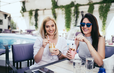 Two young and beautiful woman enjoying in private cocktail house party. They smiling and looking at camera. Swimming pool in background.