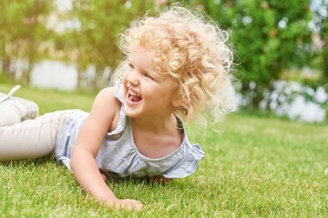Fototapeta premium Shot of a cute little girl with blonde curly angelic hair lying on the grass laughing happily looking away copyspace nature enjoyment emotions kids children family recreation summer concept.