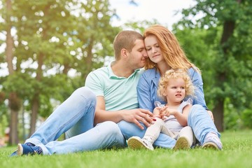 Naklejka na ściany i meble Beautiful loving couple cuddling while sitting on the grass together with their little daughter copyspace love family parenthood affection children parenting leisure holidays weekend concept.