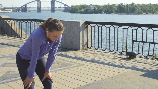 Young woman runs into the shot from left. She stops to rest for a while. Leaning her hands on the legs, she hyperventilates. Then she straightens