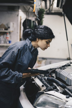 Side view of female mechanic holding digital tablet while examining car engine in auto repair shop