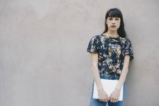 Portrait of confident female computer programmer holding laptop while standing against beige wall in office