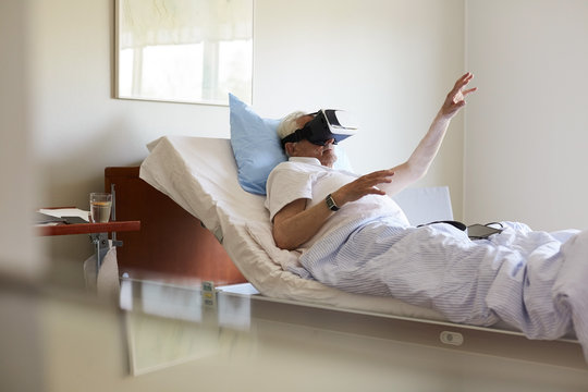 Senior man gesturing while using VR glasses on bed in hospital ward