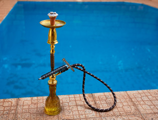 Hookah with hose and burning coal standing on a side of blue water pool.