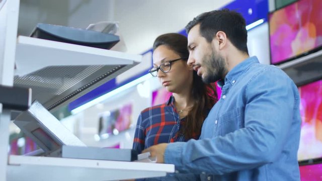Beautiful Young Couple in the Electronics Store Browsing, Looking for Newest Gadgets, Tablets and Photo/ Video Cameras Presented on the Shelves. Shot on RED EPIC-W 8K Helium Cinema Camera.