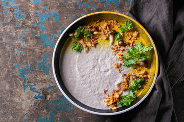 Mushroom cream soup in black ceramic bowl served with turmeric powder, forest mushrooms, greens, fried onion on black textile napkin over old dark wooden background. Top view