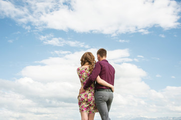 View from the back of a young couple stands in an embrace and looks out into the distance against the sky with clouds.