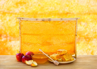 Honey in glass bowl, wooden honey dipper and honeycombs in wooden frame with full cells of honey sealed with wax and apples on wooden table on background honeycombs with full cells of honey