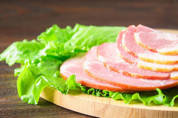 Sliced ham with fresh green lettuce leaves on a round cutting board. Meat products on a brown wooden table.