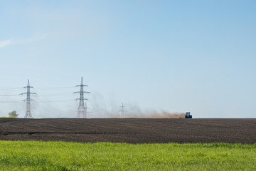 Fototapeta na wymiar blue tractor plowing field with electric power transmission pylons on background 