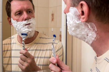 The man is going to shave standing in front of the mirror. He put a foam on his cheeks for shaving.