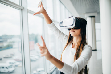 Beautiful girl using virtual reality glasses near bright window with skyscraper view outside. Business woman wearing VR goggles