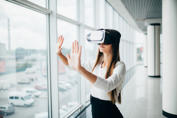 Beautiful girl using virtual reality glasses near bright window with skyscraper view outside....