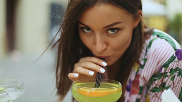 Sexy beautiful brunette woman with hair shaking by wind in dress in a city street cafe with cocktail - focus on the face and the drink, slow motion shot