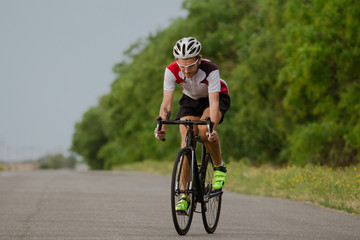 Bicycle racer in helmet and sportswear training alone on empty country road, fields and trees background   