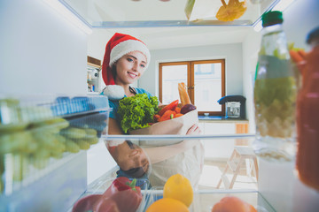 Portrait of female in santa hat standing near open fridge full of healthy food, vegetables and...