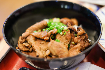 Rice with grilled pork teriyaki and spring onion on top in the black plastic bowl under the red tray for japanese food background or texture.