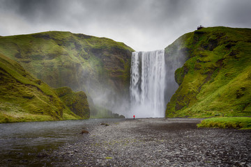 Skogafoss waterfall in Iceland. Guy in red jacket looks at the huge flow of water. Famous tourist attraction. Drammatic Sky