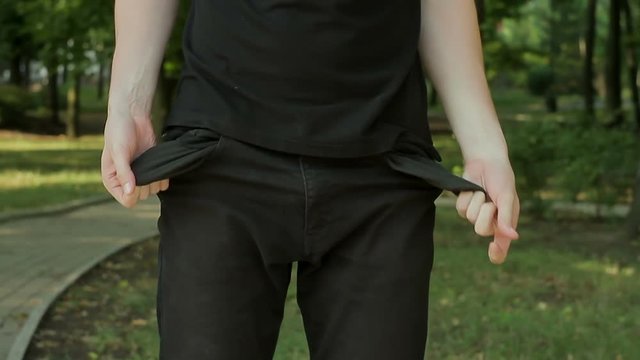 A man dressed in black standing on the nature in the park is looking for something in his pockets, puts them in his hands and turns empty pockets without money out.