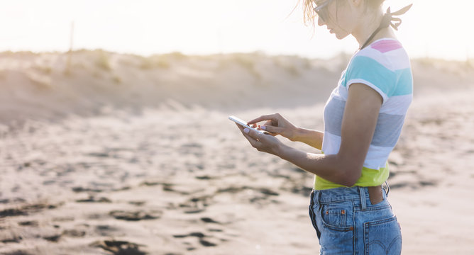 Hipster girl hold on smart phone gadget in sun sand coastline. Traveler using in female hand mobile on background beach seascape horizon flare. Tourist look on sun ocean, summer lifestyle