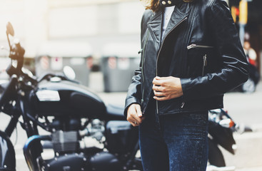 Girl unfastens black leather jacket on background motorcycle in sun flare city, hipster biker...