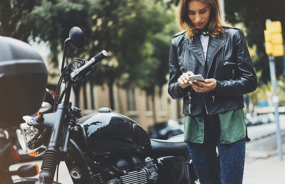 Fototapeta Girl in leather jacket holding smart phone on background motorcycle in sun flare city, hipster using in female hands and texting mobile, motorbike street lifestyle, tourist planing route in summer
