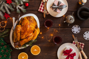 Christmas festive table is decorated with a garland. Baked chicken as a main holiday dish. Cone, dried oranges, plates, knife, fork furry spruce on a table. Top view.