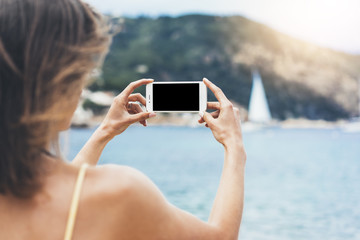 Hipster girl make photo on smart phone in coastline, mock up blank screen. Traveler using in female hand mobile on background beach seascape horizon. Tourist look on blue sun ocean, summer lifestyle