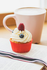 Small cupcake with raspberry and blueberry on the book, cup of hot cappuccino
