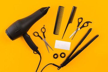 Black hair dryer, comb and scissors on yellow paper background. Top view