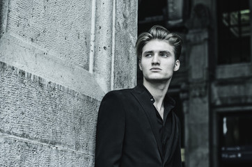Fototapeta premium Handsome blond young man, blue eyes, leaning against white, rough stone wall, looking up to a side