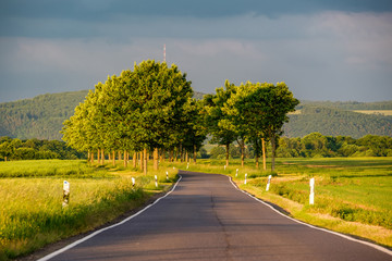 Rural road between fields in warm sunshine under dramatic sky